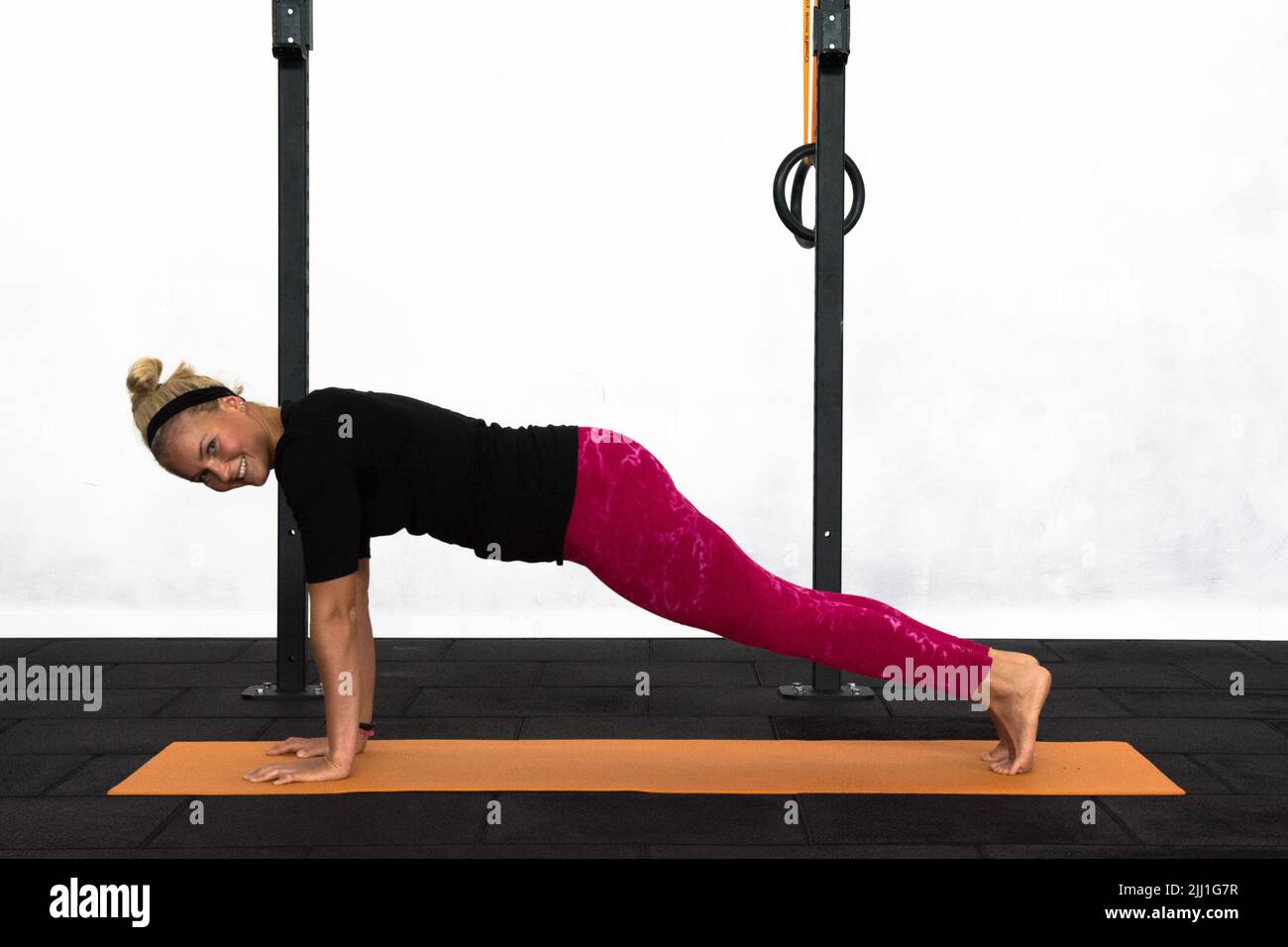 Eine blonde Jugendliche trainiert in einem Fitnessstudio mit einer Plank-Yoga-Haltung und lächelt dabei zur Kamera. `s trägt ein schwarzes Langarm-Hemd und rosa Yoga-Leggings. Stockfoto